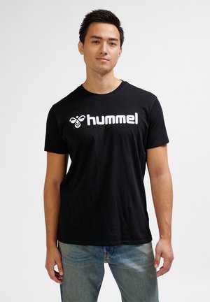 Młody mężczyzna w czarnej koszulce z logo hummel i niebieskich jeansach, stojący na jednolitym jasnym tle.