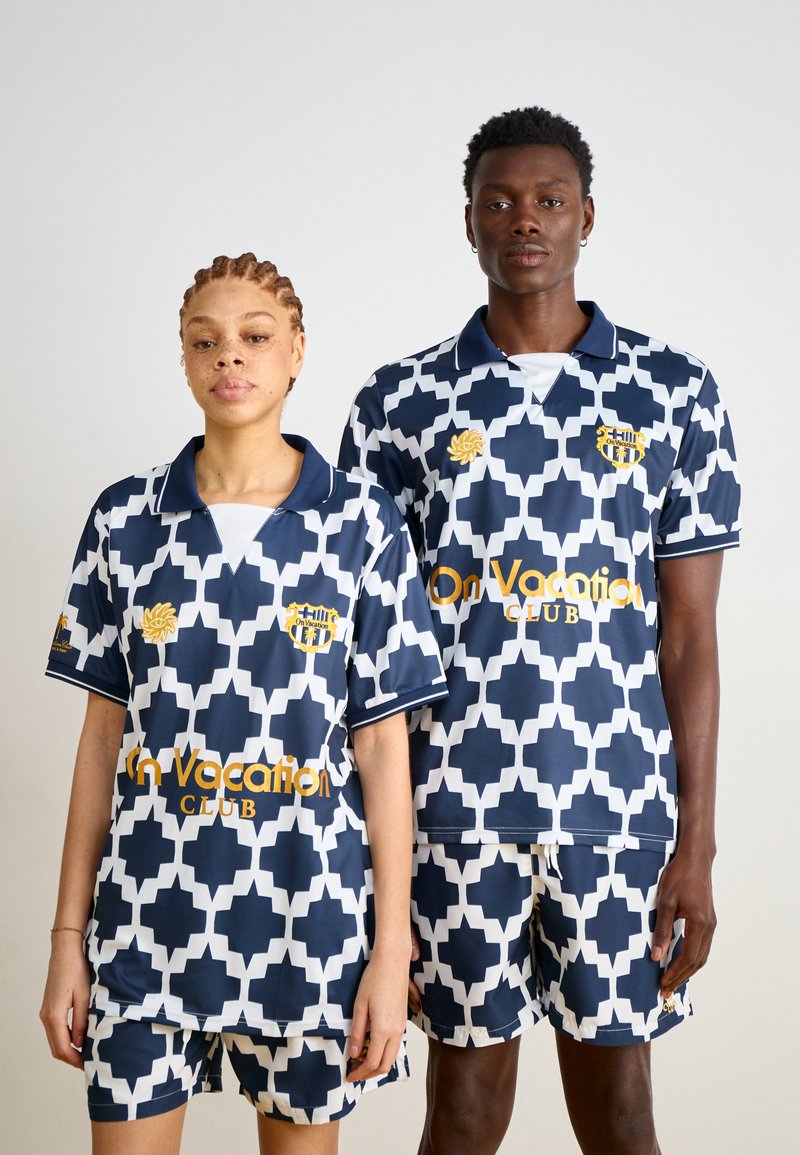 Marinblå och vit mönstrad träningskläder med geometrisk design. Inkluderar shorts och matchande kortärmade polo tröjor med logotypdetaljer.