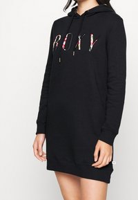 Robe noire à capuche avec inscription florale "ROXY", cordon de serrage et poignets côtelés. Texture douce et coupe décontractée, arrivant à mi-cuisse.