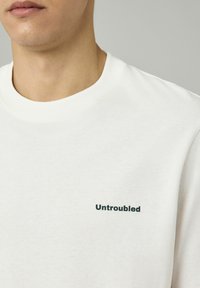T-shirt en coton blanc avec un col rond et des manches courtes, portant "Untroubled" imprimé en vert foncé sur la partie gauche de la poitrine.