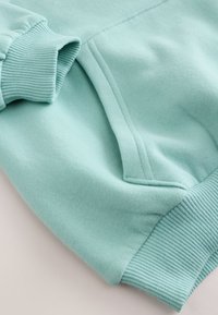 Manche de sweat-shirt bleu clair avec poignet côtelé et poche kangourou avant, montrant la texture douce du tissu en coton.