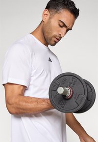 Sort vægtplade håndvægt med en sølvfarvet håndtag, holdt i en muskuløs arm. Personen bærer en tætsiddende hvid sportstrøje med en sort logo.