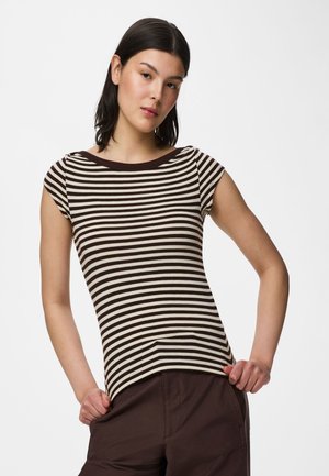 Νεαρή γυναίκα με ίσια σκούρα μαλλιά, φοράει εφαρμοστό καφέ και λευκό ριγέ κοντομάνικο πουκάμισο και σκούρο καφέ παντελόνια, τα χέρια στις τσέπες.