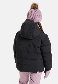 Un enfant portant une doudoune noire avec une capuche, un bonnet en tricot rose avec un pompon en fourrure, un pantalon de neige rose et des gants roses, tourné dos à l'appareil photo.