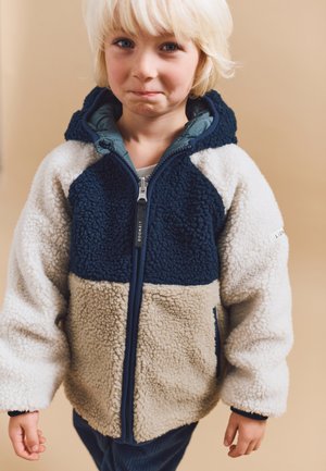 Veste en polaire avec des panneaux bleu marine, crème et beige. Elle présente une capuche, une fermeture éclair à l'avant et des poches latérales. Surface texturée avec une apparence douce.