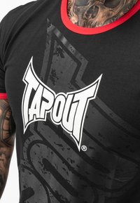 Černé tričko s červenými akcenty, vybavené výrazným bílým logem "TAPOUT" a opotřebovaným grafickým designem na látce. Krátké rukávy.
