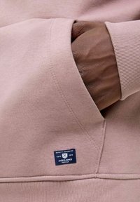 Helles rosa Sweatshirt mit einer Vorderasche, gerippten Bündchen und einem kleinen blauen Etikett mit Markendetails, das am Saum angenäht ist.