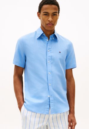 Junger Mann trägt hellblaues kurzärmeliges Hemd mit kleinem Logo und weiße Hose mit dünnen blauen vertikalen Streifen.