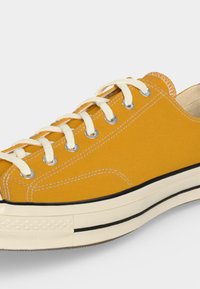 Zapatilla de lona amarilla con cordones blancos y suela blanca gruesa con rayas negras y ojales metálicos sobre un fondo liso.