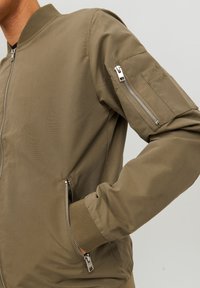 Olive green bomber jacket with a zippered front, side pockets, and an arm pocket with a zipper. Smooth fabric, ribbed collar and cuffs.