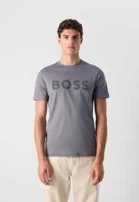 Camiseta de manga corta gris con un gran logo negro "BOSS" impreso en la parte delantera, hecha de tela suave, combinada con pantalones de color claro.