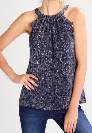 Kobieta ubrane w granatowy top na szyję bez rękawów z białym wzorem w kropki oraz niebieskie dżinsy, pozująca z jedną ręką na biodrze.