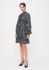 Abito floreale nero con maniche lunghe, un orlo arricciato e una vita aderente. Abbinato a décolleté nere a punta e una piccola borsa verde.