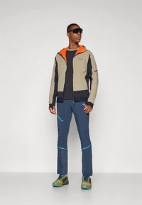 Vyras, vilkintis smėlio ir juodos spalvos švarką su oranžiniu pamušalu, tamsiai mėlynus sportinius kelnes, žalius ir mėlynus žygiams skirtus batus ir tamsius akinius nuo saulės, stovi.