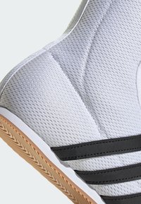 White mesh athletic shoe featuring a textured upper and black leather stripes. Gum rubber sole with a herringbone pattern for traction.
