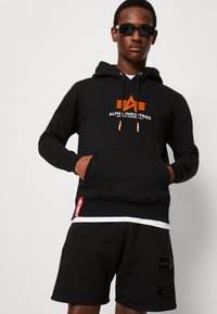 Czarna bluza z kapturem z pomarańczowym logo, sznurki oraz kieszeń z przodu. Czarne szorty z teksturowanym materiałem i detalem z logo. Akcesorium: czarne okulary przeciwsłoneczne.