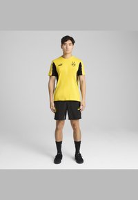 Kollane sportlik särk mustade aktsentidega, koos mustade lühikeste pükste ja Puma logo ning meeskonna embleemiga särgil. Tekstuuri välimus on sile.