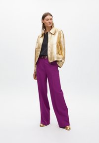 Zlatna metalik bomber jakna, crni top s točkicama, ljubičaste široke hlače i zlatne cipele. Ukupno, hrabra kombinacija boja i tekstura.