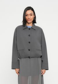 Γυναίκα που φοράει γκρι σακάκι με κουμπιά και ρυθμιζόμενο κορδόνι στη μέση πάνω από διάφανο γκρι φόρεμα, στέκεται μπροστά σε απλό λευκό φόντο.