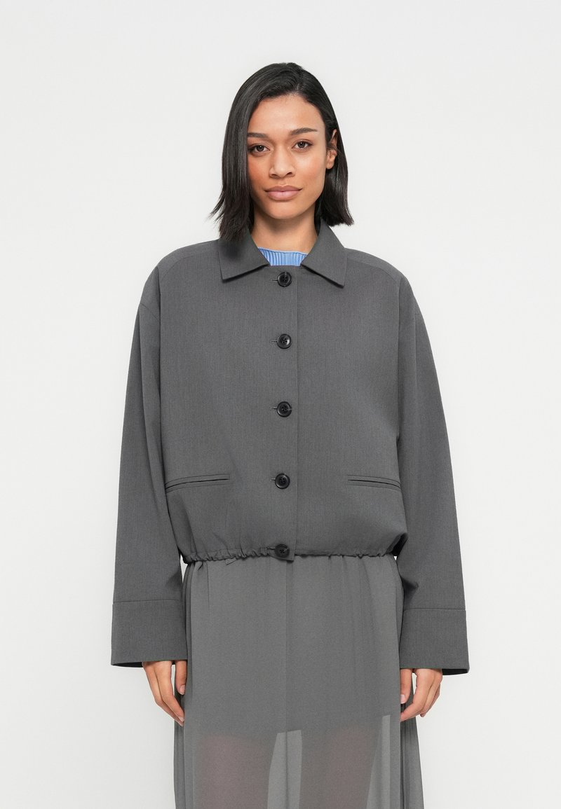 Γυναίκα που φοράει γκρι σακάκι με κουμπιά και ρυθμιζόμενο κορδόνι στη μέση πάνω από διάφανο γκρι φόρεμα, στέκεται μπροστά σε απλό λευκό φόντο.