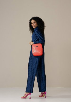 Navyblauw gestructureerd jumpsuit, roze hoge hakken, rode vierkante crossbodytas met reliëfstrepen, vastgehouden op heuphoogte, tegen een neutrale achtergrond.
