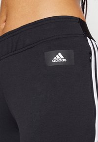 Leggings deportivos negros con una textura suave, que presentan franjas blancas a los lados y un parche con el logotipo de Adidas en la cadera.