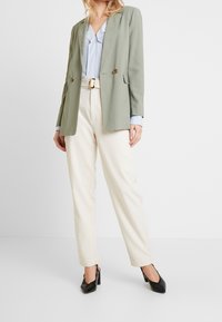 Blazer verde chiaro doppiopetto sopra una blusa azzurra; pantaloni crema; scarpe con tacco nere; tessuto liscio e vestibilità su misura.