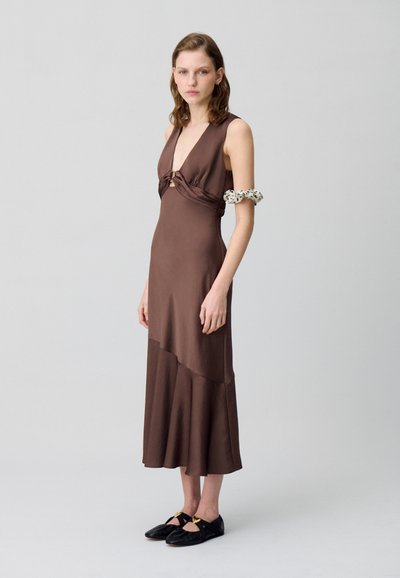 Robe sans manches marron avec un décolleté en V profond, des détails drapés et un ourlet en couches. Portée avec des chaussures noires avec des accents en or.