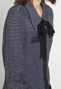 Laivastonsininen ja valkoinen houndstooth-kuvioitu takki, jossa on musta nauha yksityiskohtana kauluksessa, kiiltäviä nappeja ja teksturoitua kangasta.