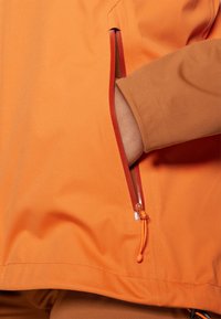 Orange outdoor jacket with a zipped pocket, contrasting red zipper, and elastic cuffs. The fabric appears smooth with a matte finish.