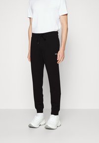 Zwarte trainingsbroek met een koord in de taille, geribde boorden en een wit "HUGO" logo op de bovenbeen, gecombineerd met witte sneakers en een basic T-shirt.