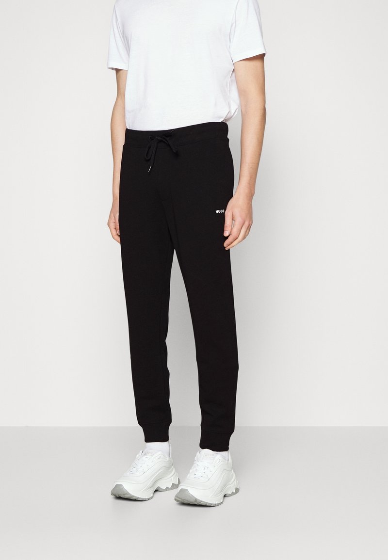 Zwarte trainingsbroek met een koord in de taille, geribde boorden en een wit "HUGO" logo op de bovenbeen, gecombineerd met witte sneakers en een basic T-shirt.