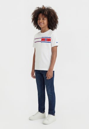 Enfant aux cheveux bouclés portant un t-shirt de sport blanc avec des rayures rouges et bleues, un jean foncé et des baskets blanches, souriant et debout.