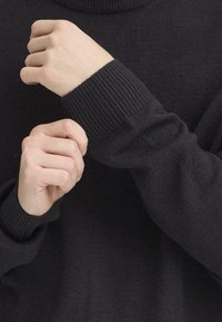 Maglione grigio scuro con polsini a costine, morbida texture e vestibilità rilassata. Messa a fuoco sulle mani che regolano la manica. Design minimalistico, senza loghi visibili.