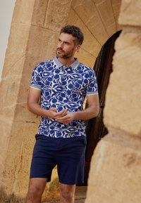 Korte mouwen blauw-wit gestreept polo shirt met een grijze kraag, gecombineerd met donkerblauwe shorts. Het ontwerp bevat abstracte vormen.