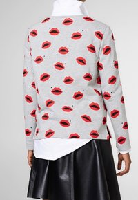 Grå sweater med gentaget grafisk mønster af røde læber. Har en sideslids og ribbede manchetter. Bæret over en hvid top og sort nederdel.