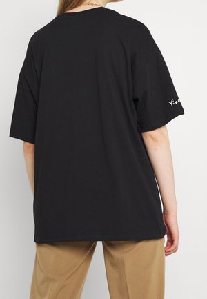 Henkilö, jolla on päällään löysä musta T-paita, jossa on teksti "Yind" oikeassa hihassa, ja beiget housut, näkyvissä takaa valkoista taustaa vasten.