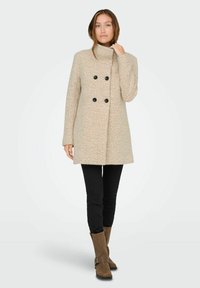 Manteau beige à double boutonnage avec des boutons noirs, col montant et manches longues, porté sur un pantalon noir ajuste et des bottes marron jusqu'aux genoux.