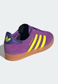 Fioletowy zamszowy sneaker z żółtymi paskami i logo, gumowa podeszwa, zaokrąglony nosek oraz teksturowane akcenty na pięcie i języku.