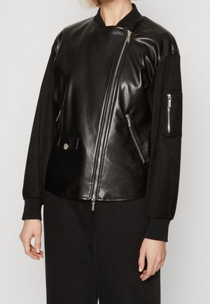 Osoba ubrana w czarną asymetryczną skórzaną kurtkę zapinaną na zamek, z kieszeniami na zamek i ściągaczami na rękawach, w połączeniu z czarnymi spodniami.