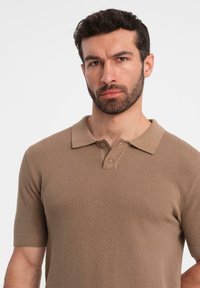 Camisa polo de punto beige con mangas cortas, que presenta un patrón texturizado y una tapeta de tres botones en el cuello.