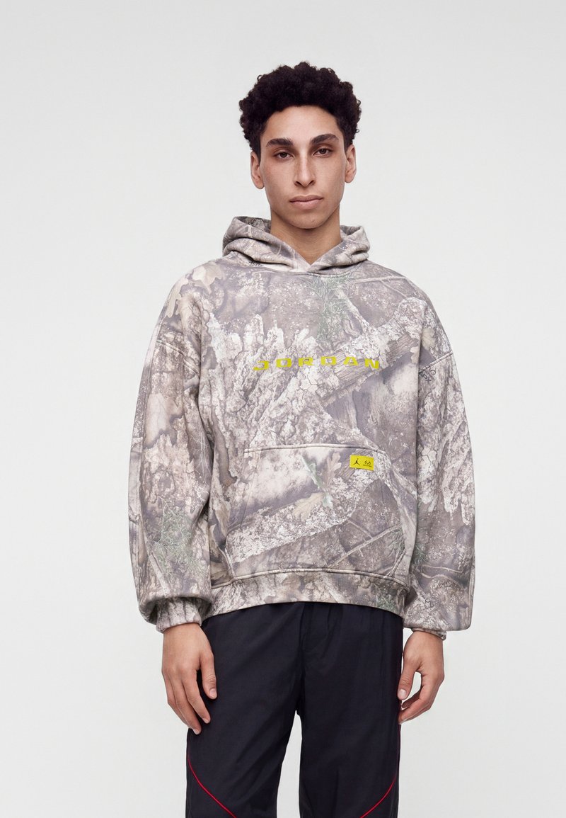Jeune homme portant un sweat à capuche Jordan camouflage gris avec un logo jaune et un pantalon noir avec des détails rouges, debout sur un fond blanc uni.