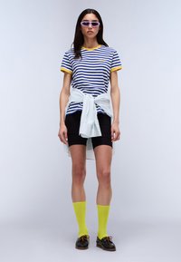 Stripet skjorte i blå og hvit med gul kant, kombinert med svarte shorts og knelange neon gule sokker. Hvit strikkegenser knyttet rundt livet.