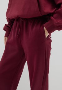 Pantalones de chándal burdeos fabricados en un tejido suave, con una cinturilla elástica con cordón, bolsillos laterales y puños acanalados.