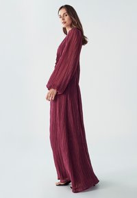 Dlouhé splývavé šaty v tmavě burgundské barvě s texturovaným, skládacím designem. Mají dlouhé rukávy, zapínání na knoflíky vpředu a volný střih.