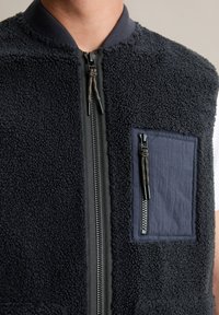 Fleece prsluk tamnoplave boje, s punim prednjim zatvaračem, rebrastim ovratnikom i džepom na prsima sa zatvaračem s kontrastnim materijalom.