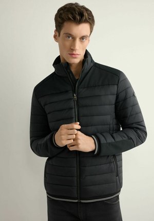 Młody mężczyzna w czarnej, pikowanej kurtce zapinanej na suwak z wysokim kołnierzem, stojący na tle jasnego, jednolitego tła, ręce poprawiające suwak na wysokości klatki piersiowej.