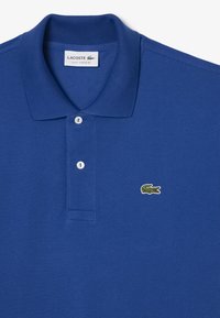 Kirkkaan sininen polo-paitaa, jossa on kaksinappinen kaulus ja vihreä brodeerattu krokotiililogo vasemmalla rinnalla.