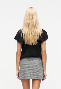 Ξανθιά γυναίκα με μακριά κυματιστά μαλλιά, φορώντας μαύρο τοπ με κοντά μανίκια και μίνι φούστα καρό σε μαύρο και άσπρο, όρθια με την πλάτη γυρισμένη.
