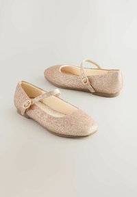 Rozā zelta mirdzoši Mary Jane apavi ar mazu sprādzi un bēšiem iekšzoliem, novietoti uz vienkāršas gaiši pelēkas virsmas.